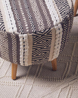 A grey and white patterned stool with a rounded top and wooden legs, placed on a textured light-colored rug. The fabric features various geometric designs in shades of grey, cream, and white