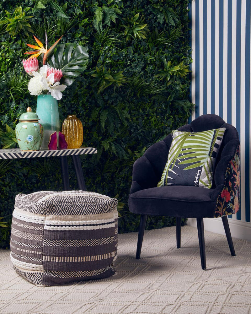A grey and white pouffe sits on a tiled floor beside a black armchair with a colorful cushion. A small side table holds decorative vases and a vibrant floral arrangement, with a green wall of plants in the background