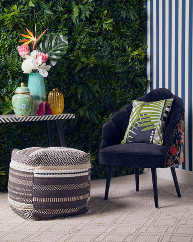 A grey and white pouffe sits on a tiled floor beside a black armchair with a colorful cushion. A small side table holds decorative vases and a vibrant floral arrangement, with a green wall of plants in the background