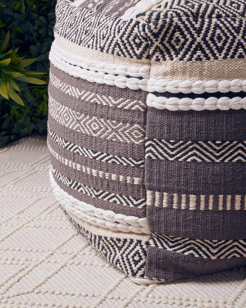 A round pouffe featuring a textured design in shades of grey and white, with various woven patterns and stripes. The pouffe is placed on a light-colored surface, surrounded by greenery in the background