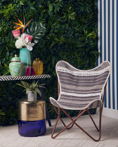 A grey and white butterfly chair with a sleek design is positioned next to a small table. The table features a colorful arrangement of decorative items, including vases and a potted plant, set against a backdrop of lush green foliage and striped wallpaper