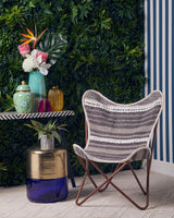 A grey and white butterfly chair with a sleek design is positioned next to a small table. The table features a colorful arrangement of decorative items, including vases and a potted plant, set against a backdrop of lush green foliage and striped wallpaper
