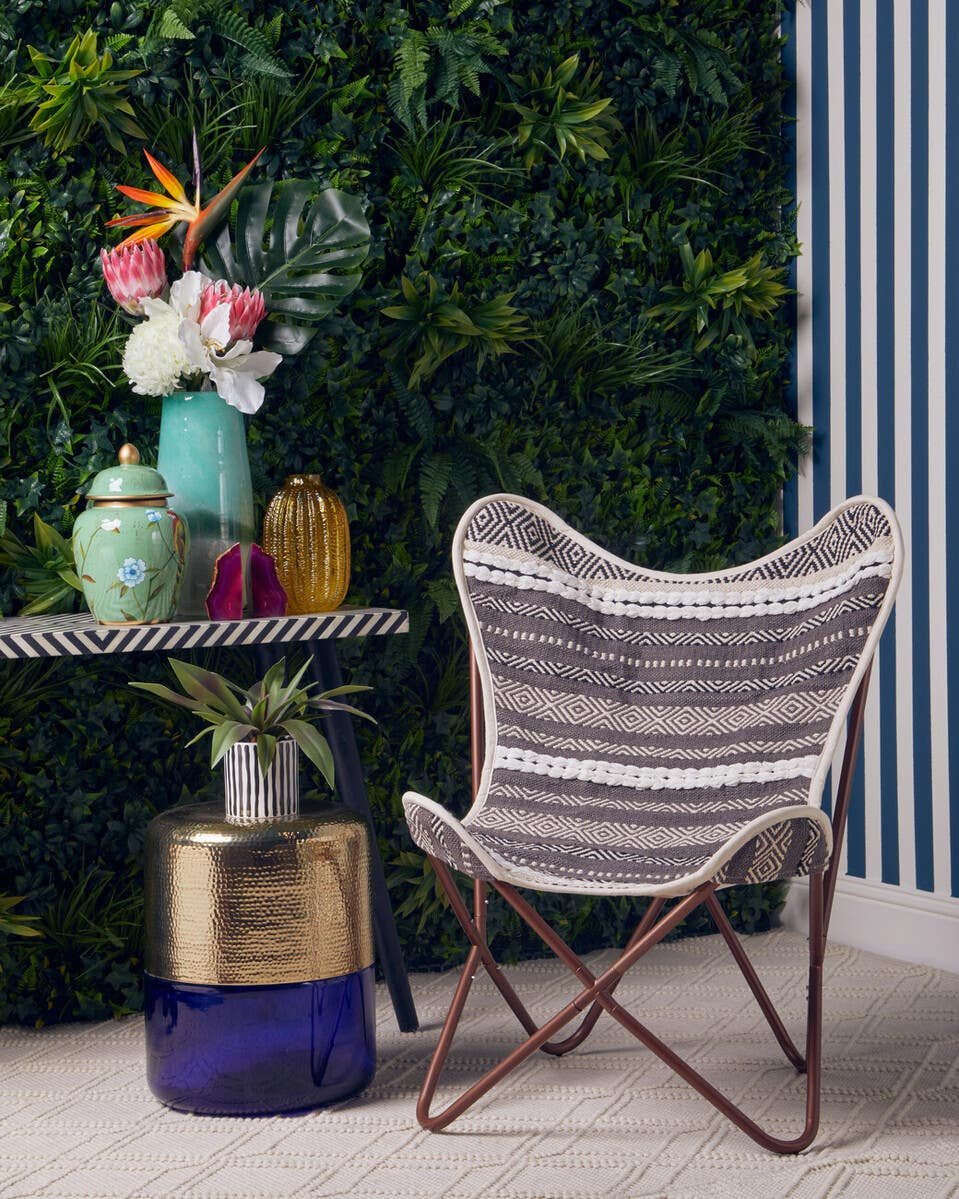 A grey and white butterfly chair with a sleek design is positioned next to a small table. The table features a colorful arrangement of decorative items, including vases and a potted plant, set against a backdrop of lush green foliage and striped wallpaper