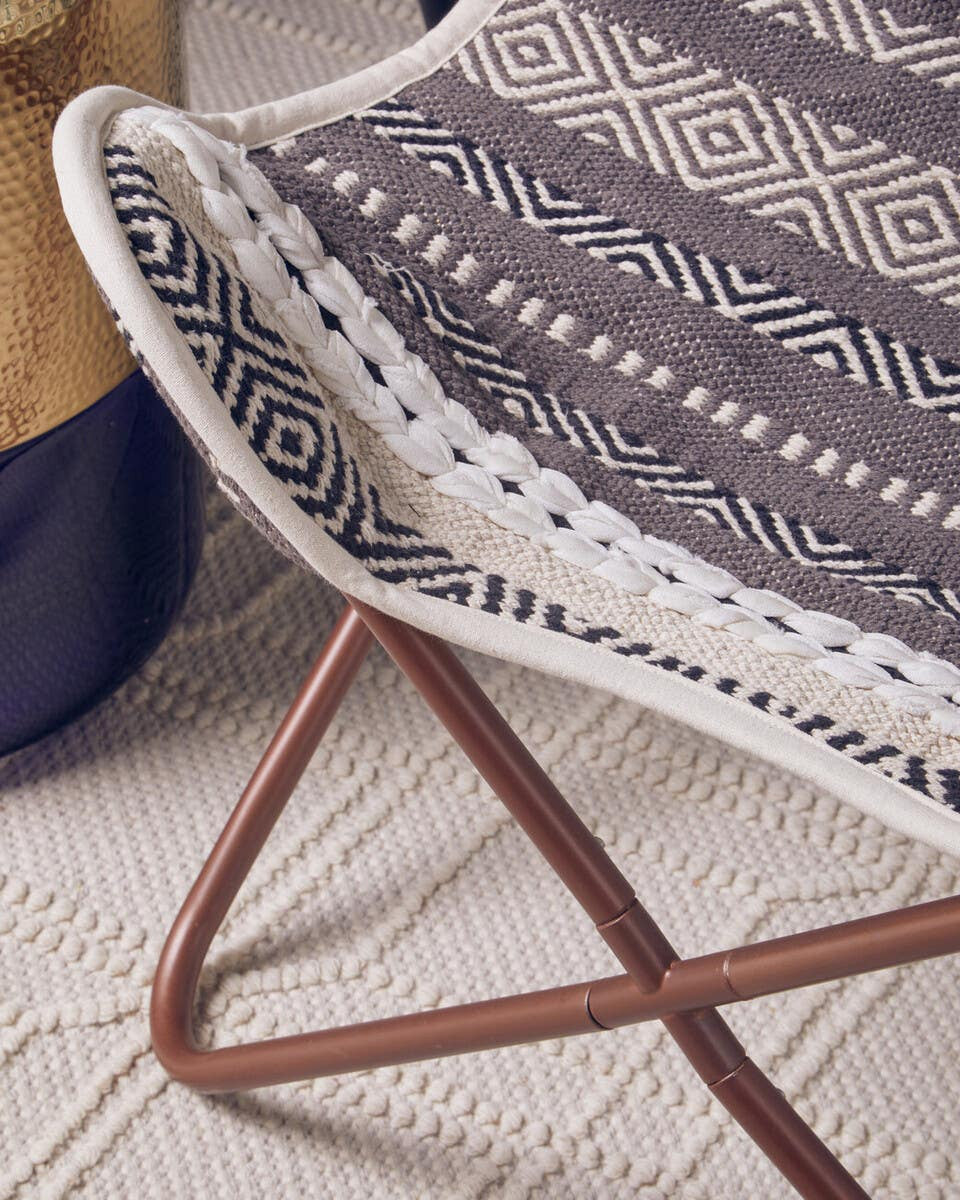 Grey and white butterfly chair featuring a woven seat with a geometric pattern and a sturdy metal frame in a warm brown finish, placed on a textured light-colored rug