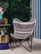 Grey and white butterfly chair with a distinctive winged design, featuring a woven fabric seat and a sturdy metal frame. The chair is positioned on a light-colored floor, surrounded by greenery and colorful decor elements in the background