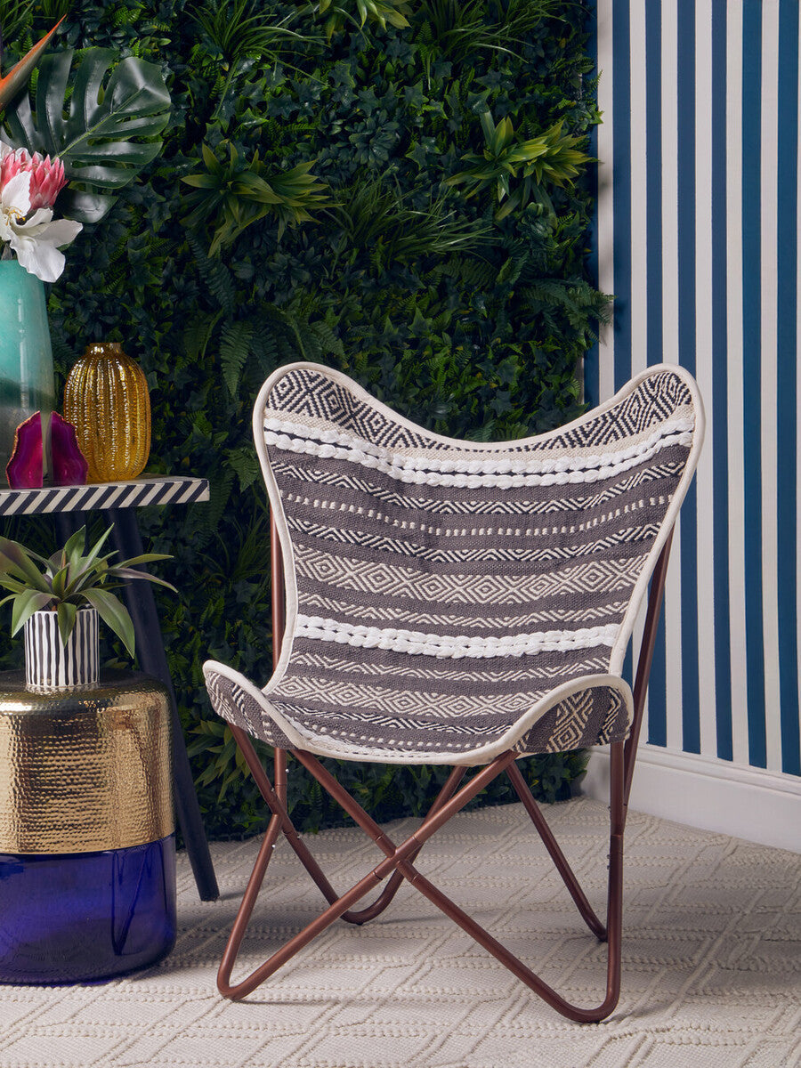 Grey and white butterfly chair with a distinctive winged design, featuring a woven fabric seat and a sturdy metal frame. The chair is positioned on a light-colored floor, surrounded by greenery and colorful decor elements in the background