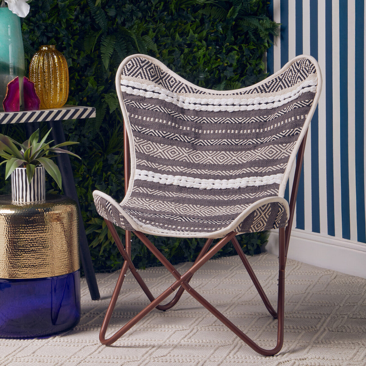 A grey and white butterfly chair with a curved, woven seat design, supported by a sleek, brown metal frame. The chair is positioned on a light-colored rug, with a decorative plant and a small table in the background, featuring striped blue and white walls