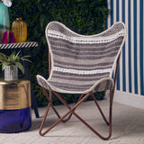A grey and white butterfly chair with a curved, woven seat design, supported by a sleek, brown metal frame. The chair is positioned on a light-colored rug, with a decorative plant and a small table in the background, featuring striped blue and white walls
