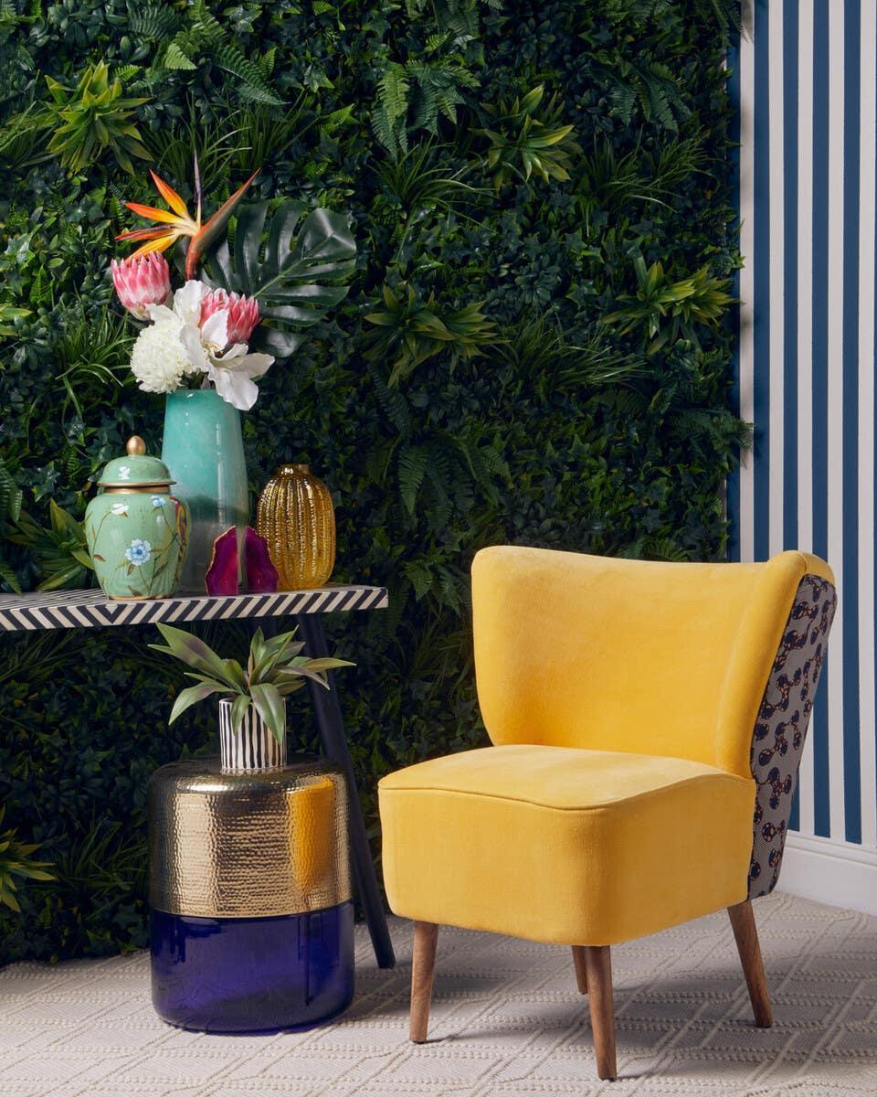Yellow accent chair with patterned side, set against green plant wall and striped wallpaper