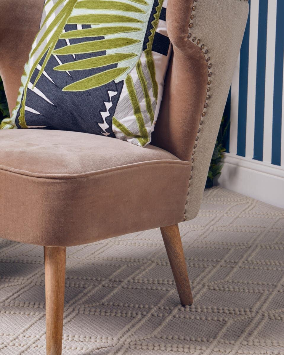 Beige velvet accent chair with green leaf cushion and wooden legs on patterned carpet