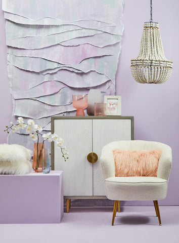 Cream and grey two-door cabinet made of mango wood, featuring a round gold handle, placed against a textured light purple wall. A white armchair with a pink cushion sits beside the cabinet, and a decorative pendant light hangs above. A small decorative vase and a bowl are positioned on top of the cabinet