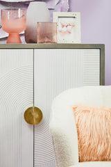Cream and grey two-door cabinet made of mango wood, featuring a sleek design with a round gold handle. The cabinet is placed against a pastel purple wall, with decorative items on top, including a pink glass vase, two pink glasses, and a book with a white cover. A soft, light-colored cushion is partially visible beside the cabinet