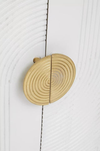 Close-up view of a round, gold-colored cabinet handle with a textured design, mounted on a cream and grey wooden cabinet door. The wood features a subtle grain pattern, enhancing the natural look of the furniture