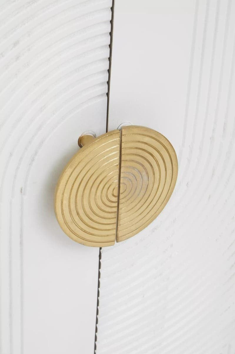 Close-up view of a round, gold-colored cabinet handle with a textured design, mounted on a cream and grey wooden cabinet door. The wood features a subtle grain pattern, enhancing the natural look of the furniture