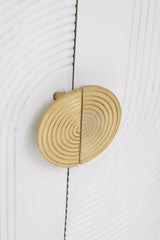 Close-up view of a round, gold-colored cabinet handle with a textured design, mounted on a cream and grey wooden cabinet door. The wood features a subtle grain pattern, enhancing the natural look of the furniture