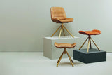 Buffalo tan leather footstool with a sleek design, featuring a cushioned top and wooden tripod legs, positioned alongside a matching chair and another footstool on a light-colored floor against a neutral backdrop