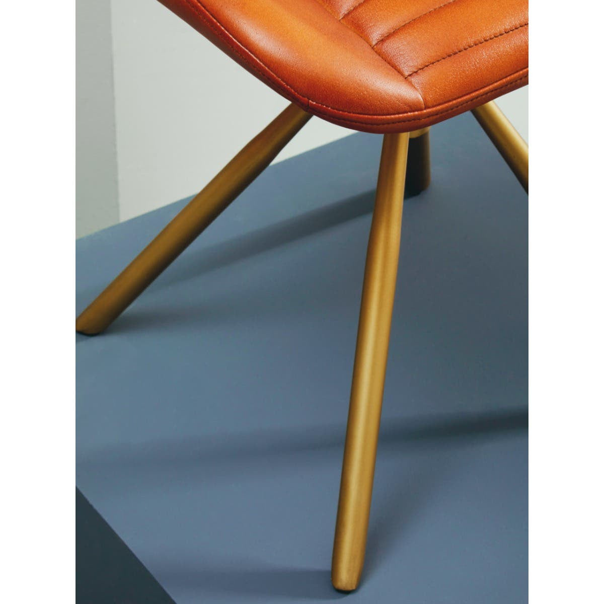 Buffalo tan leather footstool with a quilted design on the seat, supported by four wooden legs. The footstool is positioned on a blue surface, highlighting its warm color and modern aesthetic