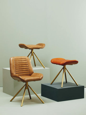 Buffalo tan leather footstool with a cushioned top and wooden tripod legs, positioned on a black square pedestal, surrounded by two matching tan leather chairs on white pedestals against a light-colored wall