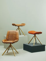 Buffalo tan leather footstool with a smooth surface and a minimalist design, featuring wooden legs that provide a sturdy base. The footstool is positioned on a black platform, alongside two matching tan leather chairs with similar wooden legs, set against a light-colored wall