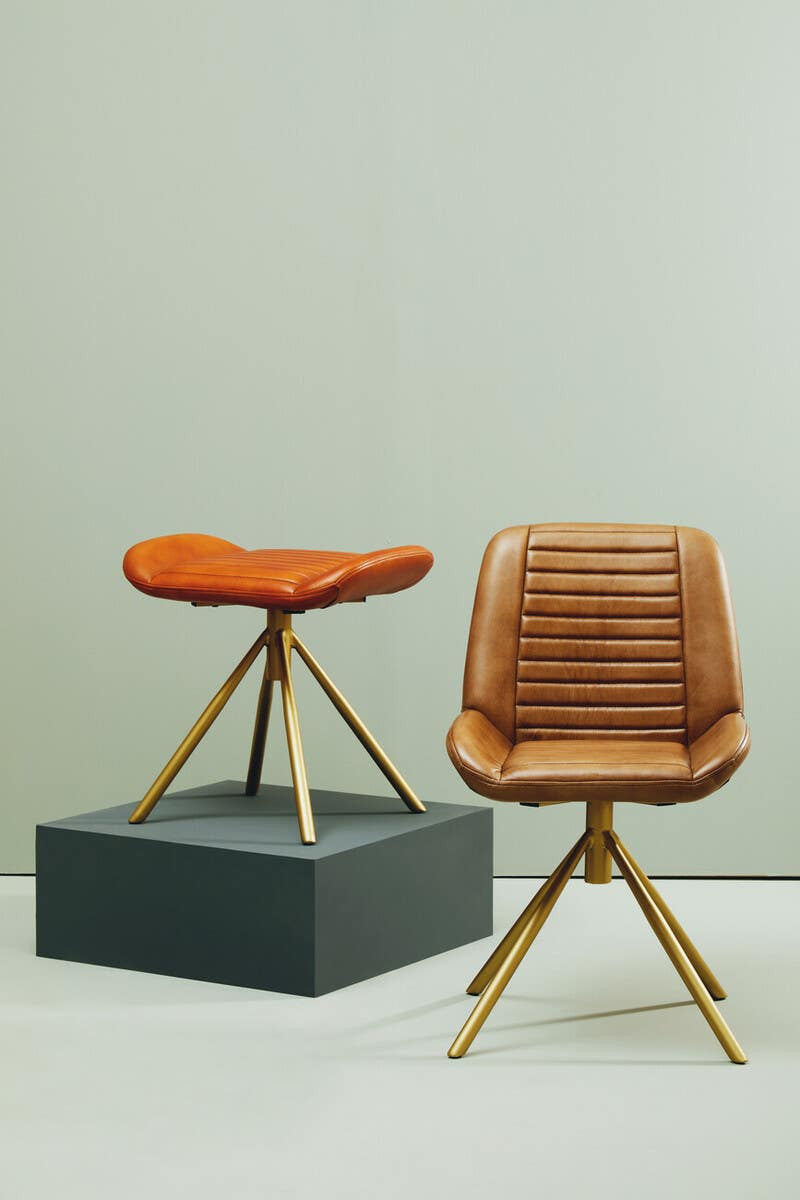 Buffalo tan leather footstool with a curved seat, positioned on a black pedestal, alongside a matching chair featuring a textured seat and wooden legs, set against a light-colored wall
