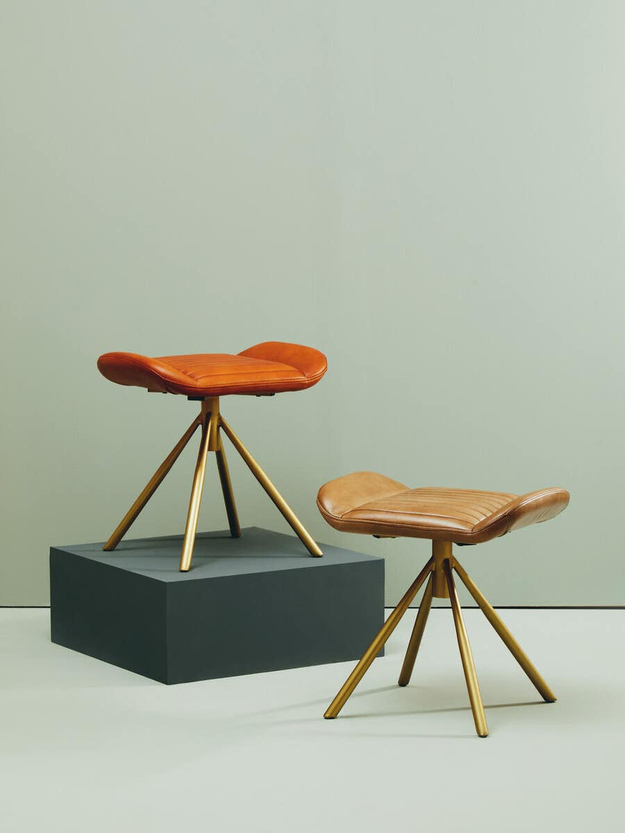 Buffalo tan leather footstool with a curved seat and wooden tripod legs, positioned on a dark gray platform against a light-colored wall
