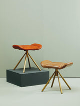 Buffalo tan leather footstool with a curved seat and wooden tripod legs, positioned on a dark gray platform against a light-colored wall