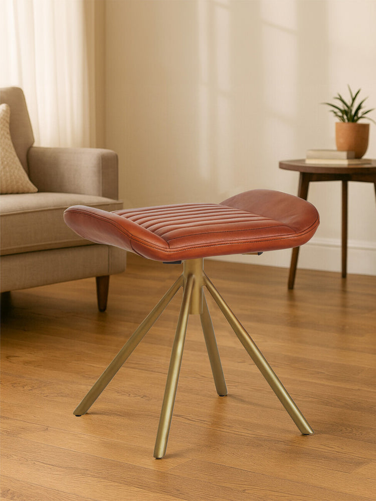 Buffalo tan leather footstool with a curved top and three slender, angled metal legs, placed on a wooden floor in a well-lit room with a sofa and a side table in the background