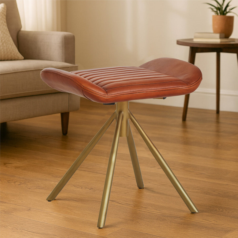 Buffalo tan leather footstool with a curved top and a sleek, modern design, supported by three gold metal legs, placed on a wooden floor in a cozy living room setting. A light-colored sofa and a potted plant are visible in the background