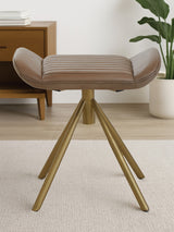 Buffalo grey leather footstool with a curved top and wooden tripod legs, positioned on a light-colored floor in a modern living space. A wooden side table and a plant are visible in the background