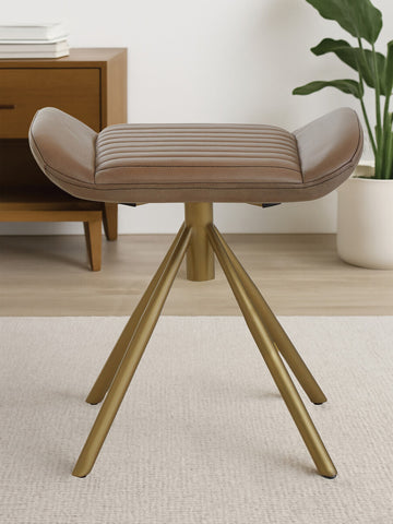 Buffalo grey leather footstool with a cushioned top and natural wood tripod legs, placed on a light-colored rug in a modern living space. A wooden side table and a plant are visible in the background