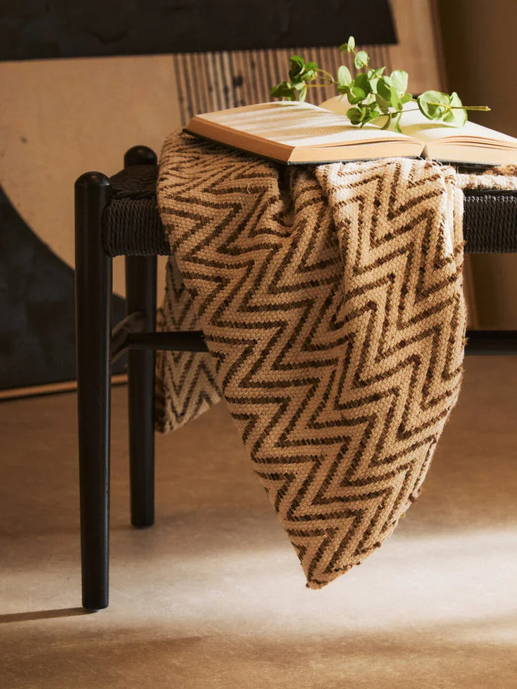 Brown zigzag throw blanket draped over black bench with open book and green plant