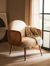 Cream fabric armchair with wooden legs and rattan sides, throw blanket draped on seat