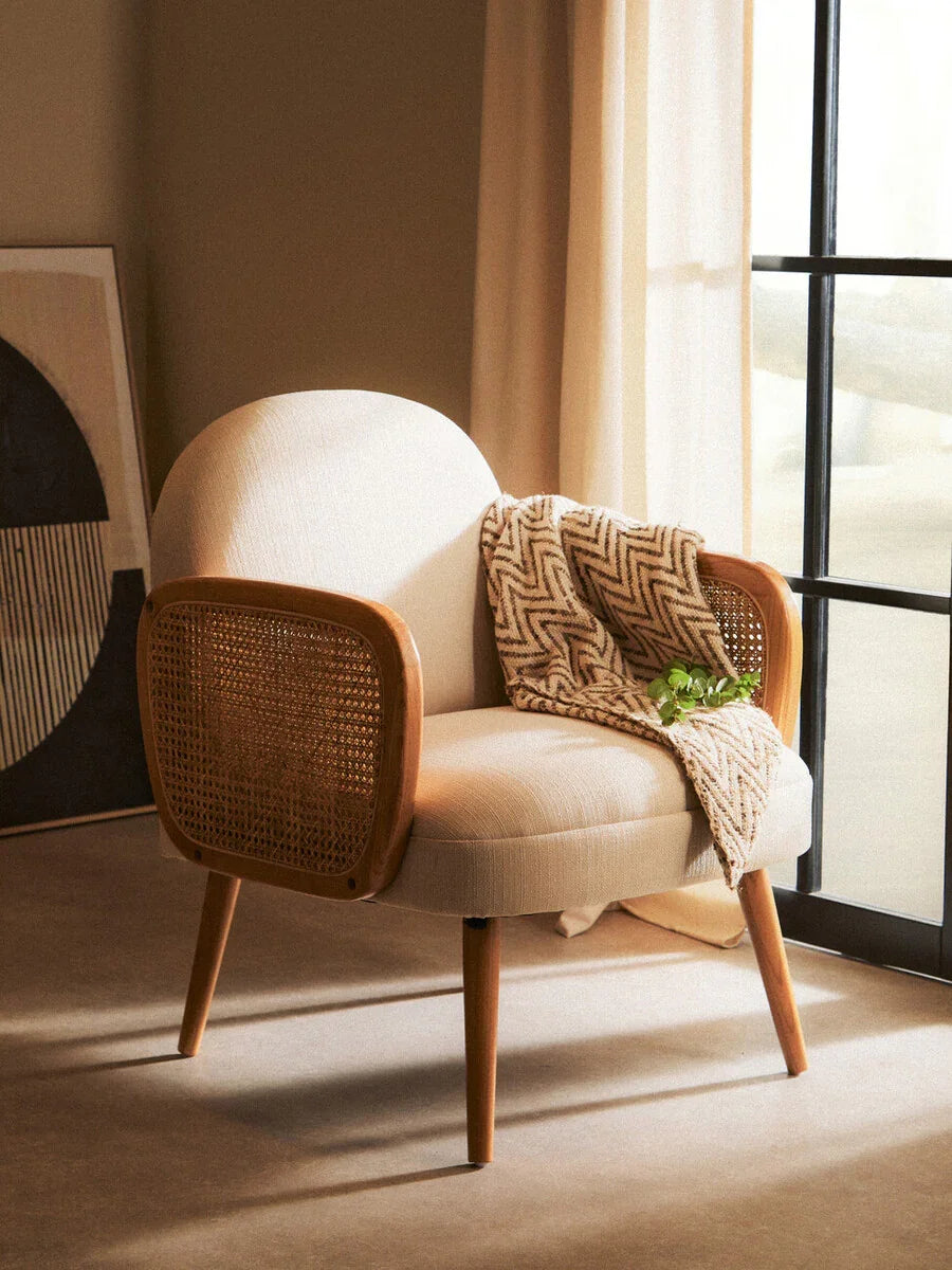 Cream fabric armchair with wooden legs and rattan sides, throw blanket draped on seat
