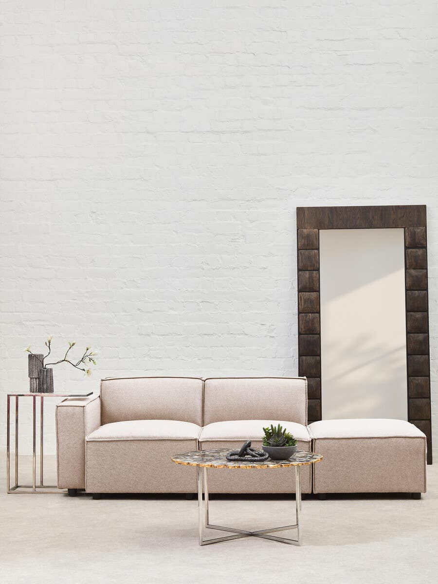 Armless modular sofa in natural fabric, featuring a minimalist design with soft cushions and a light beige color. Positioned next to a modern side table with a decorative vase and a small plant, alongside a large mirror reflecting the room's light. The background consists of a textured white wall
