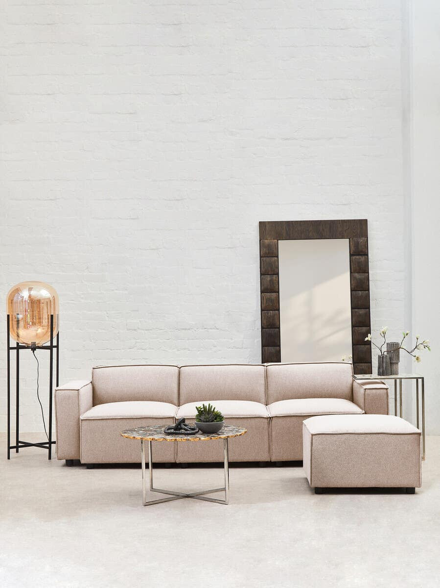 Armless modular sofa in natural fabric, featuring a light beige color, positioned in a bright room with a large mirror and a decorative lamp on a side table. A small green plant is placed on the sofa, complementing the modern aesthetic