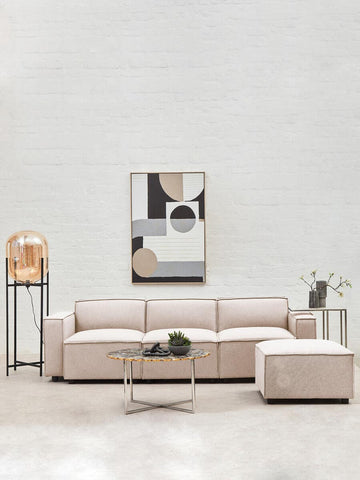 A modern armless modular sofa in a soft natural fabric, featuring a light beige color. The sofa is arranged in an L-shape with a matching ottoman, set against a neutral wall. A stylish coffee table with a decorative plant sits in front, and a contemporary artwork is displayed above the sofa. A floor lamp and a small side table are also visible in the scene