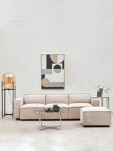 A modern armless modular sofa in a soft natural fabric, featuring a light beige color. The sofa is arranged in an L-shape with a matching ottoman, set against a neutral wall. A stylish coffee table with a decorative plant sits in front, and a contemporary artwork is displayed above the sofa. A floor lamp and a small side table are also visible in the scene