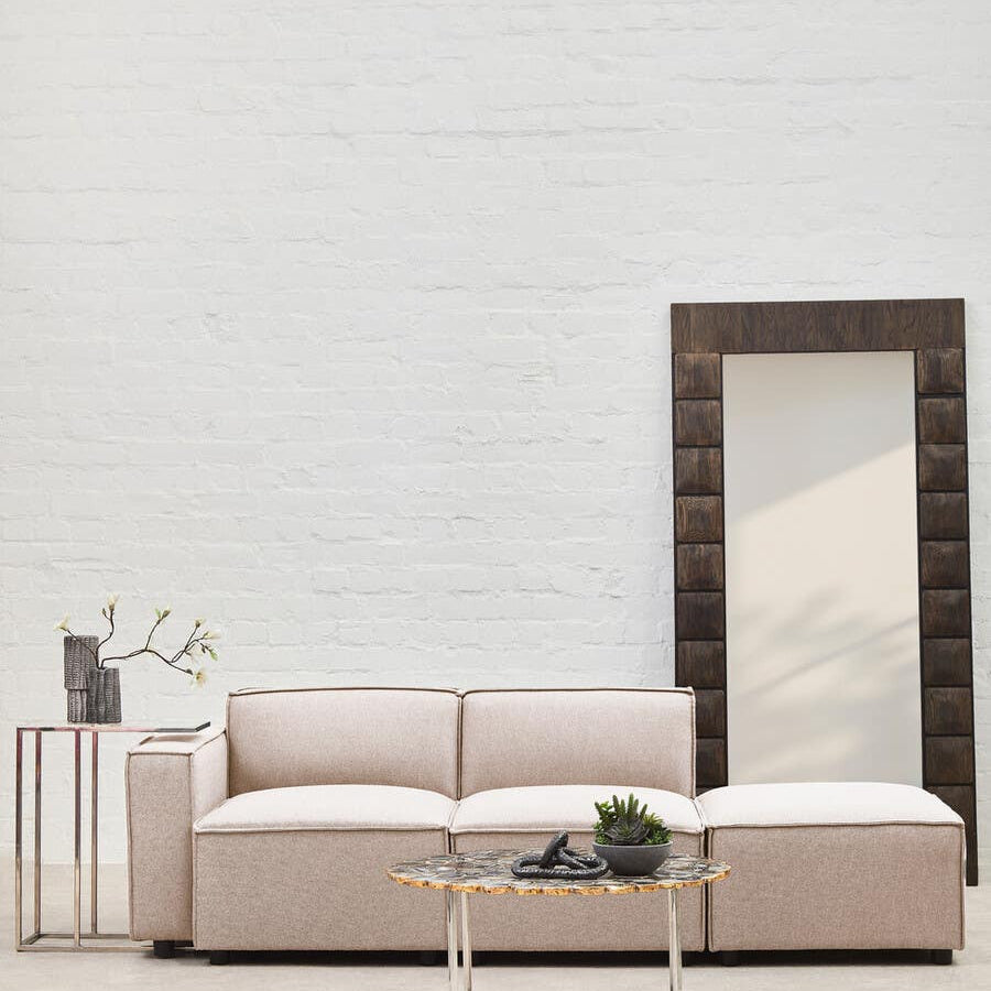 Modular ottoman upholstered in natural fabric, featuring a minimalist design with clean lines, positioned next to a sleek side table and a large decorative mirror against a light-colored wall