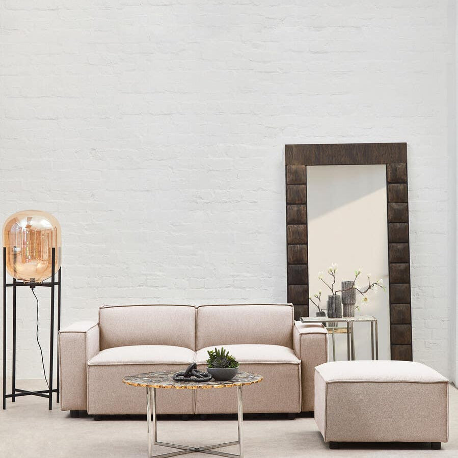 A modular ottoman in natural fabric sits beside a light-colored sofa in a modern living room. The space features a large mirror on the wall, a decorative lamp on a stand, and a small plant on the coffee table, all set against a neutral backdrop