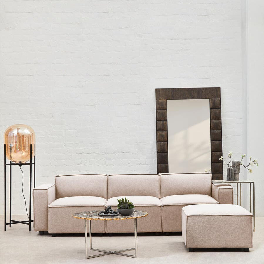 A modular ottoman upholstered in natural fabric, positioned next to a light-colored sectional sofa. The setting features a tall mirror and a decorative lamp on a sleek side table, all against a neutral wall. A small plant is placed on the ottoman, adding a touch of greenery to the arrangement