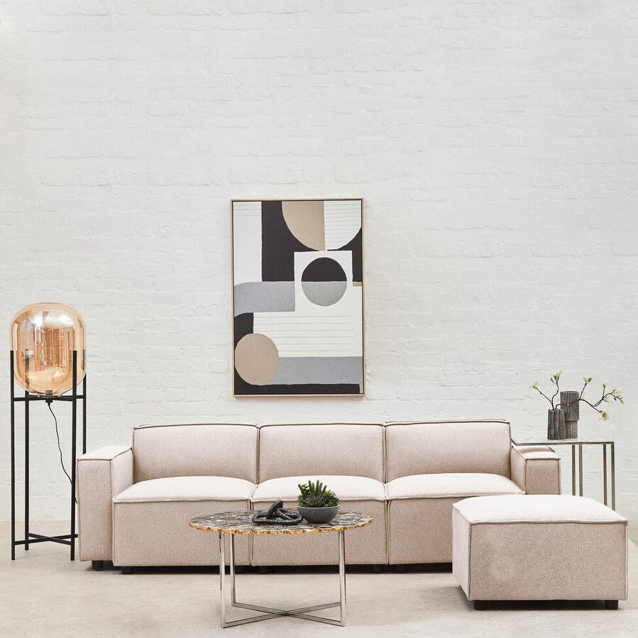 A modular ottoman upholstered in natural fabric, positioned next to a light-colored sectional sofa. The ottoman features a clean, contemporary design and is complemented by a glass coffee table and decorative items, including a plant and a lamp. A large abstract artwork hangs on the wall behind the furniture, enhancing the modern aesthetic of the space
