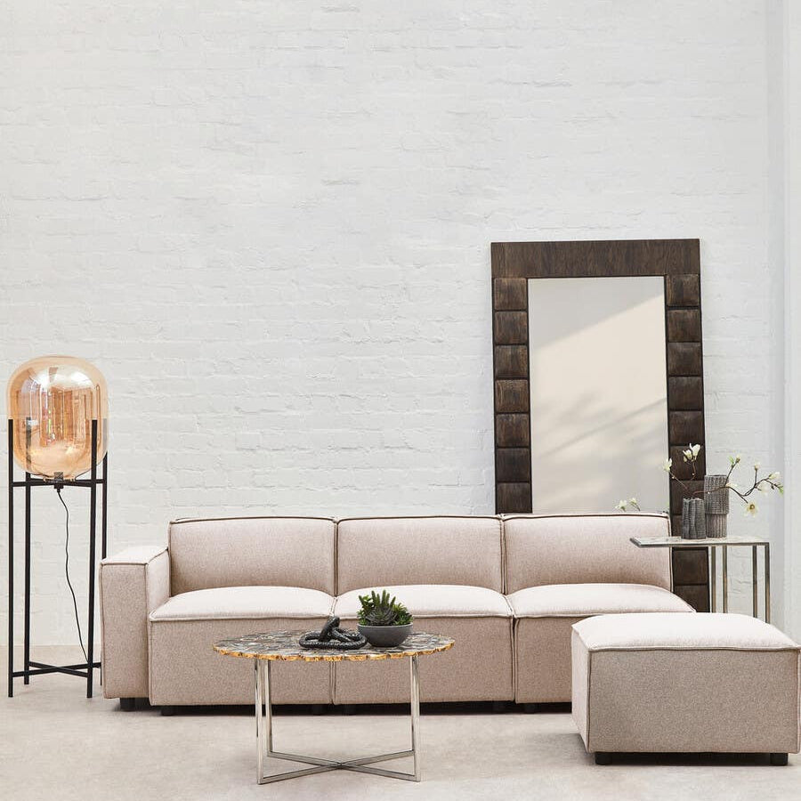 A modular ottoman upholstered in natural fabric, featuring a clean and modern design. It is positioned next to a light-colored sectional sofa, with a minimalist coffee table in front. A large mirror and a decorative lamp are visible in the background against a neutral wall. The flooring is light wood, enhancing the airy feel of the space