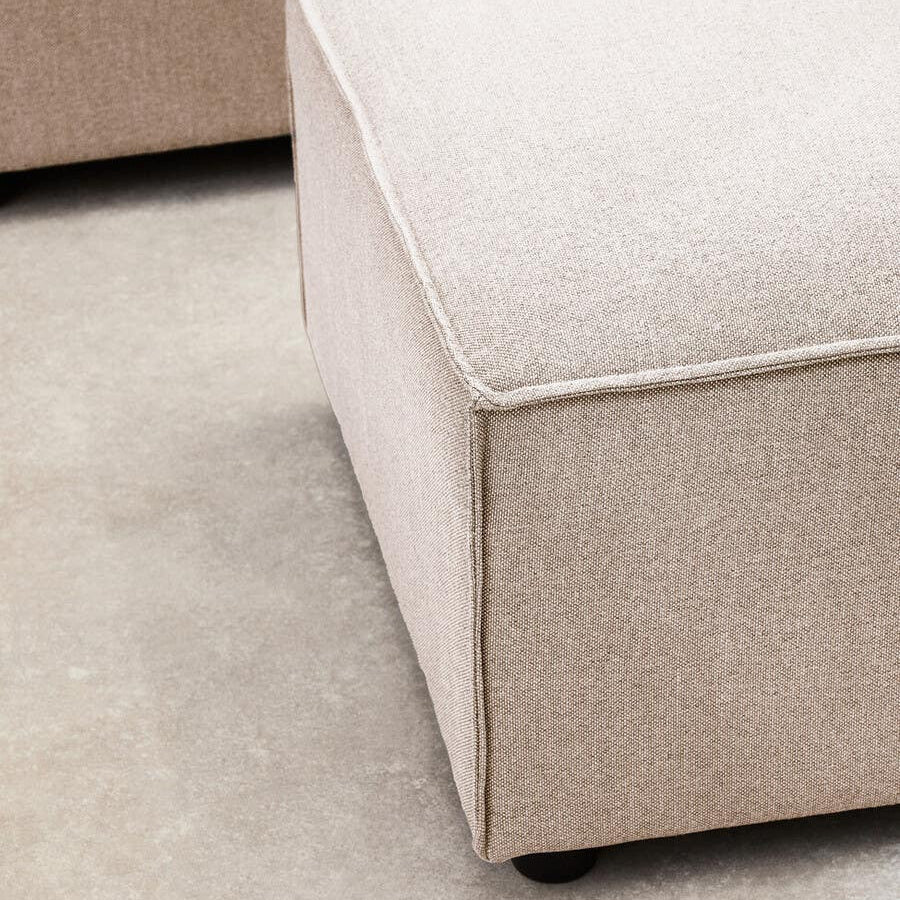 A corner view of a modular ottoman upholstered in natural fabric, featuring a soft, square top and clean lines. The ottoman has a light beige color with a subtle texture, resting on a smooth surface