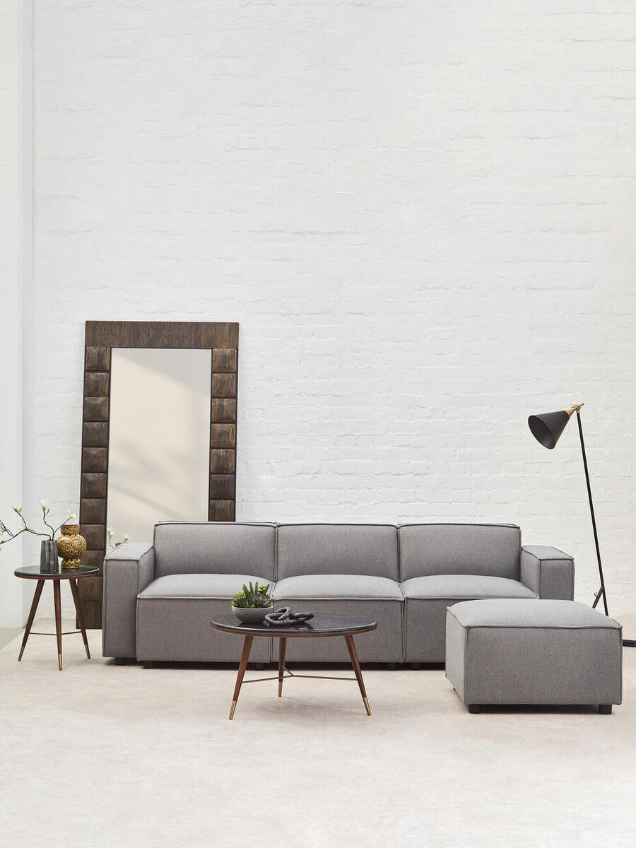 A modern grey fabric modular sofa with a right arm, featuring clean lines and plush cushions, positioned in a bright room with white walls. A large mirror with a decorative frame stands beside the sofa, and a small side table with a lamp is placed nearby. A matching ottoman is positioned in front of the sofa on a light-colored rug