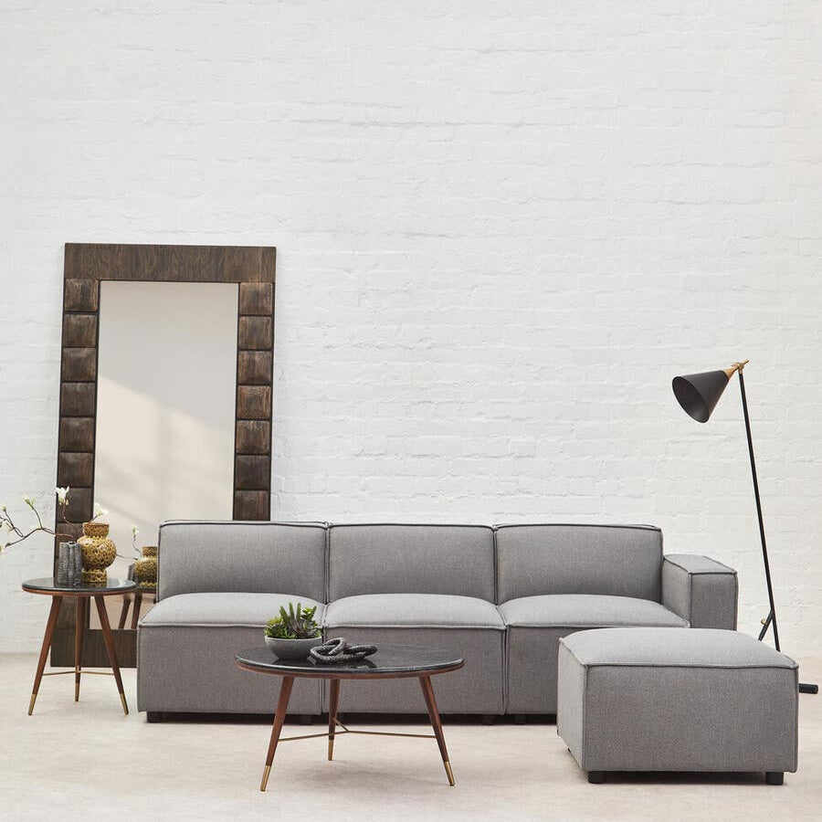 A grey fabric right arm modular sofa with a contemporary design, featuring plush cushions and a sleek silhouette. The sofa is positioned in a bright room with a large mirror and a stylish floor lamp nearby, complemented by a small side table. The flooring is light-colored, enhancing the modern aesthetic of the space