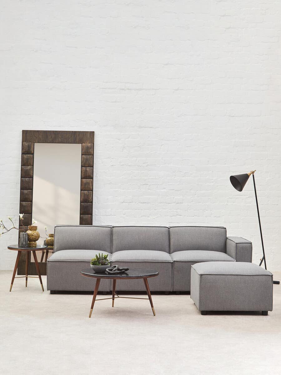 A grey fabric right arm modular sofa with a contemporary design, featuring plush cushions and a sleek silhouette. The sofa is positioned in a bright room with a large mirror and a stylish floor lamp nearby, complemented by a small side table. The flooring is light-colored, enhancing the modern aesthetic of the space