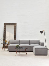 A grey fabric right arm modular sofa with a contemporary design, featuring plush cushions and a sleek silhouette. The sofa is positioned in a bright room with a large mirror and a stylish floor lamp nearby, complemented by a small side table. The flooring is light-colored, enhancing the modern aesthetic of the space