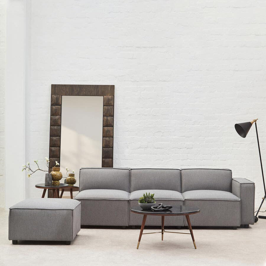 Grey fabric right arm modular sofa with plush cushions, accompanied by a matching ottoman, set against a light-colored wall. A large mirror and a floor lamp are visible in the background, adding to the modern decor