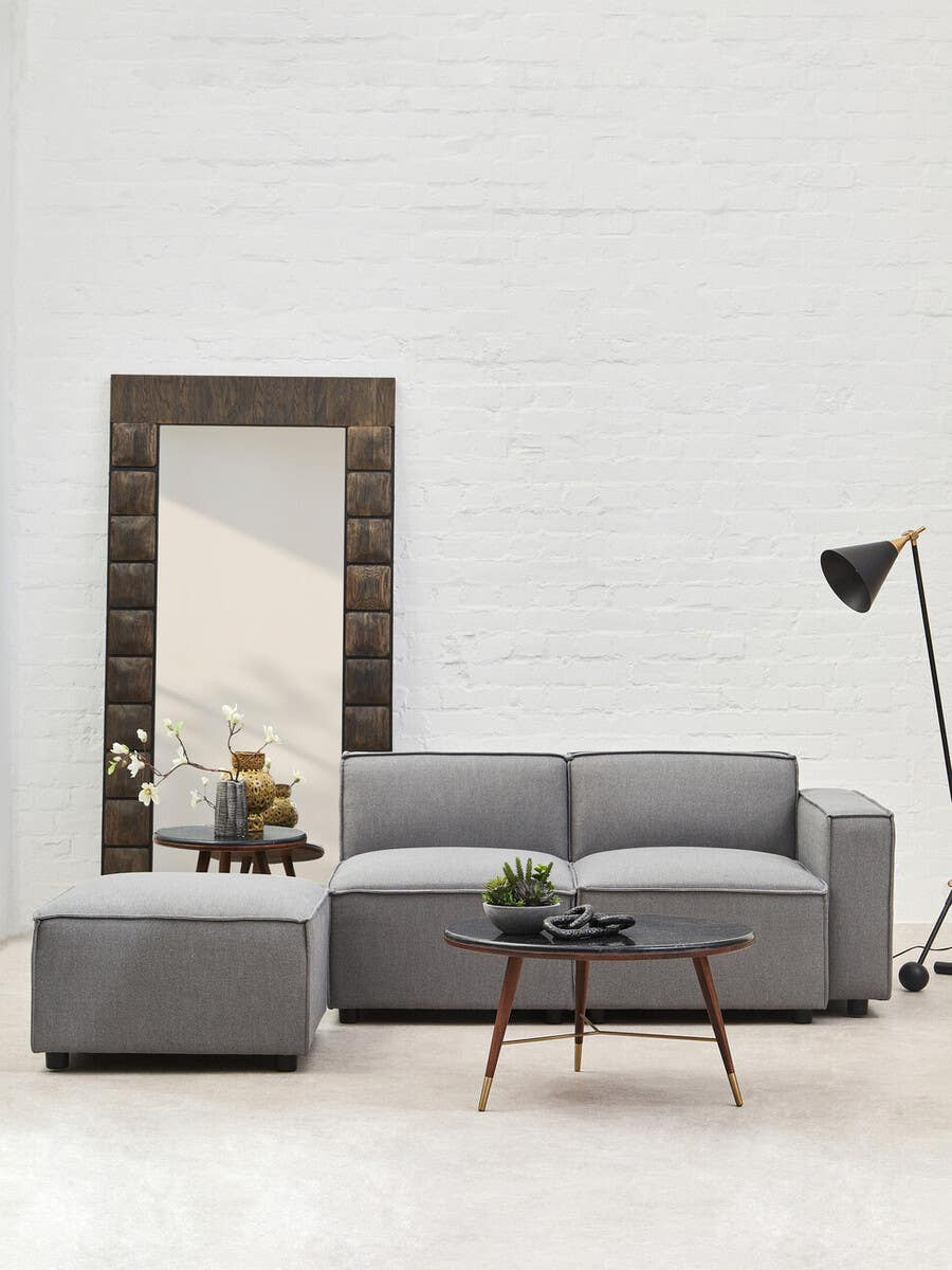 Grey fabric right arm modular sofa with plush cushions, accompanied by a matching ottoman, set against a white wall. A large mirror and a floor lamp are visible in the background, along with a small wooden coffee table featuring decorative items