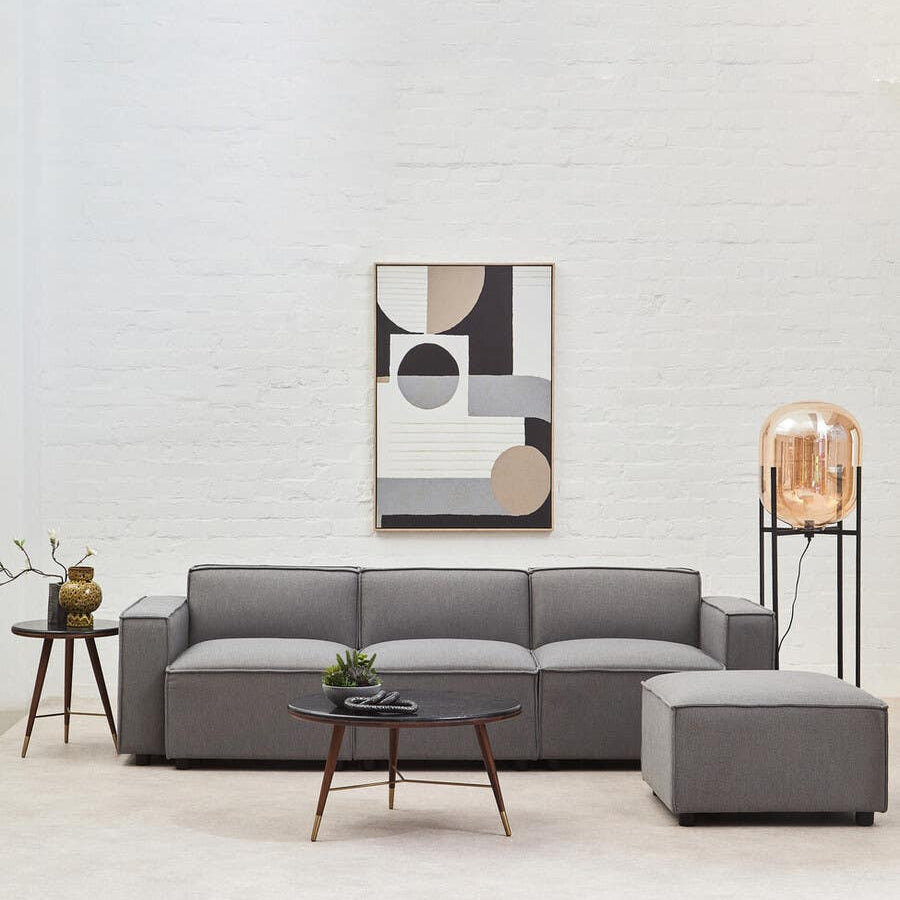 Grey fabric right arm modular sofa with a modern design, featuring clean lines and a low profile. The sofa is positioned in a bright room with a light-colored wall, accompanied by a small round coffee table and a decorative plant. A piece of abstract wall art hangs above the sofa, adding a contemporary touch to the space